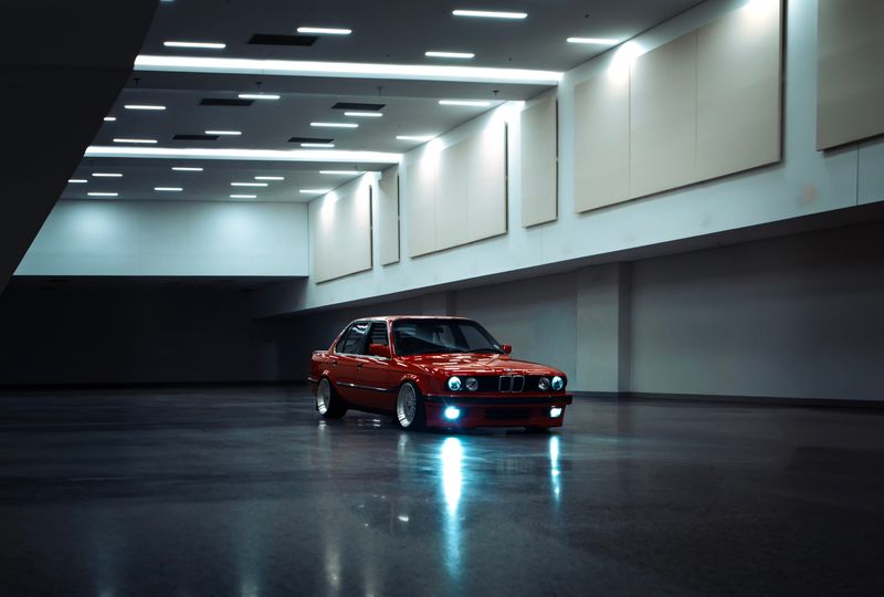 A red BMW E30 alone in a wide, empty underground parking garage, mirrored in the wet floor beneath bright ceiling strips.