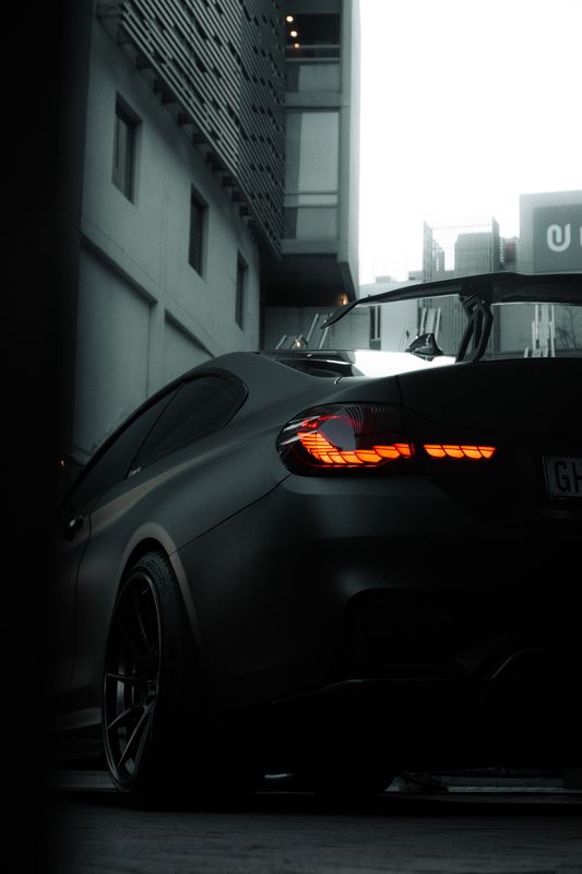 The rear quarter of a silver BMW M4 beneath a concrete overhang lit from above by orange strip lights.