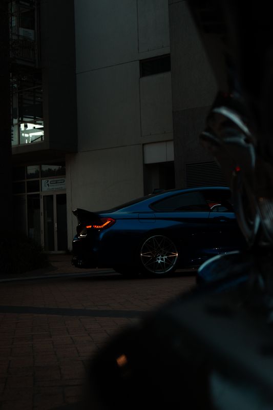 A blue BMW M4 parked on a quiet, unlit street at dusk, seen past a dark shape in the foreground.