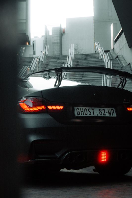 A black BMW M4 with a tall rear wing, its tail lights glowing, parked in a dim concrete garage beneath a stairwell.