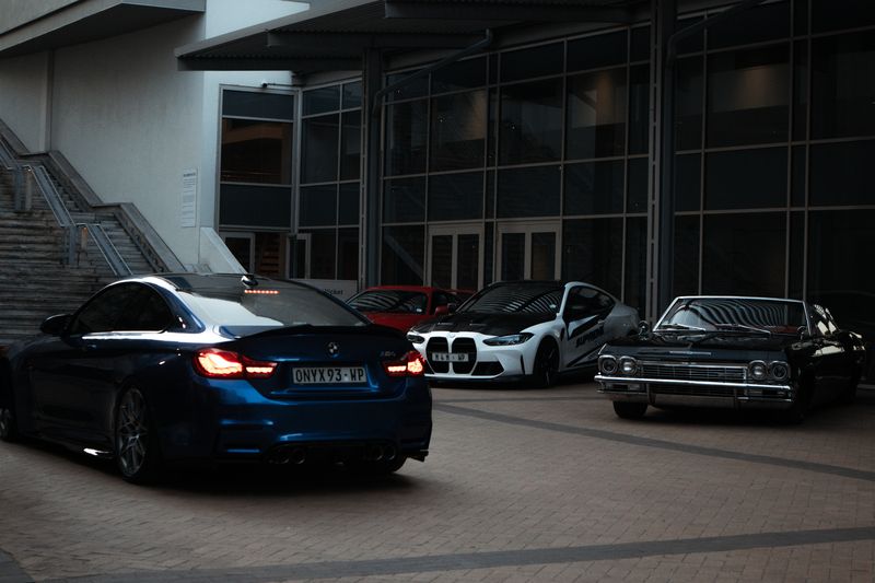 Four cars drawn up in a paved courtyard: a blue BMW M4 in front, then two BMWs and a dark classic parked side by side.
