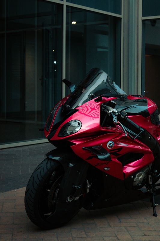 A red Ducati sports motorcycle standing on paving against a wall of brick piers and dark glass.
