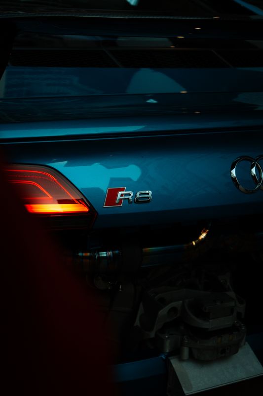 The rear of a blue Audi R8 in a garage, its tail light lit, with kit bags and tools stacked to one side.