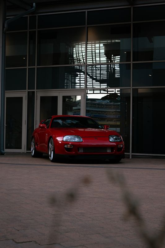 A red Toyota Supra Mk4 standing on paving before a glass-fronted building, its shape repeated across the dark window panes.