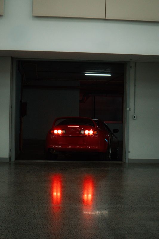 The rear of a red car standing in a lit garage doorway, its tail lights reflected in wet concrete.